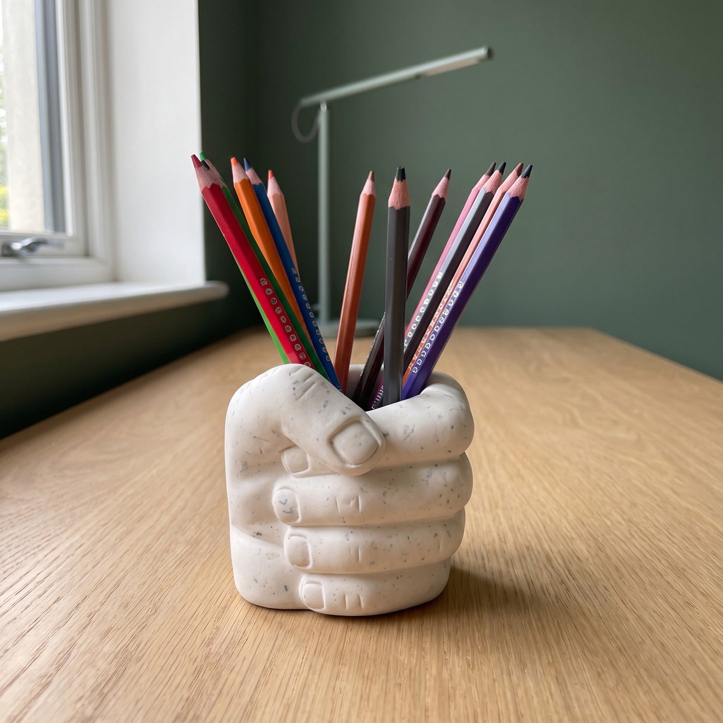 Pot à crayons Main rempli de crayons sur un bureau en bois, devant un mur vert et une lampe contemporaine