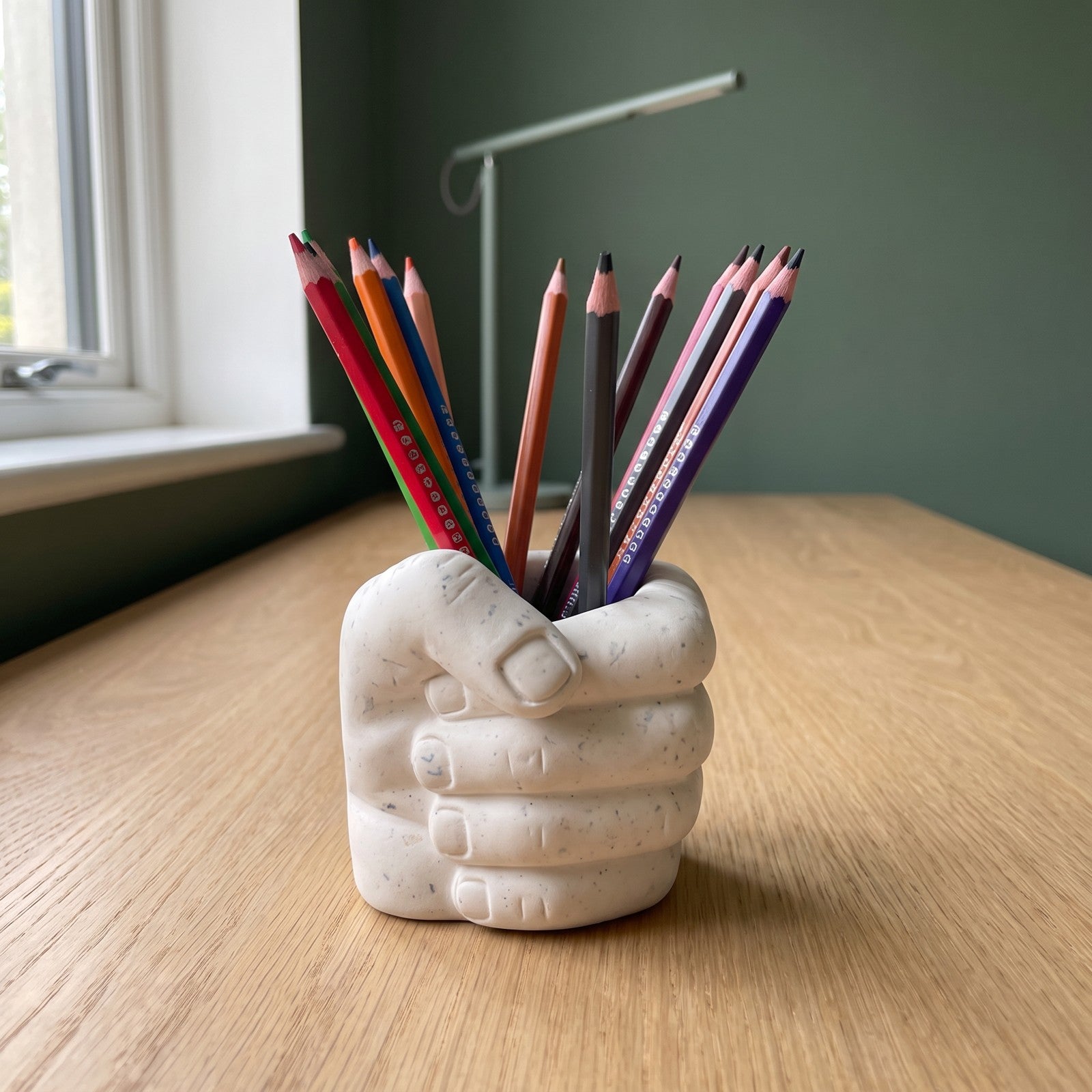 Pot à crayons Main rempli de crayons sur un bureau en bois, devant un mur vert et une lampe contemporaine